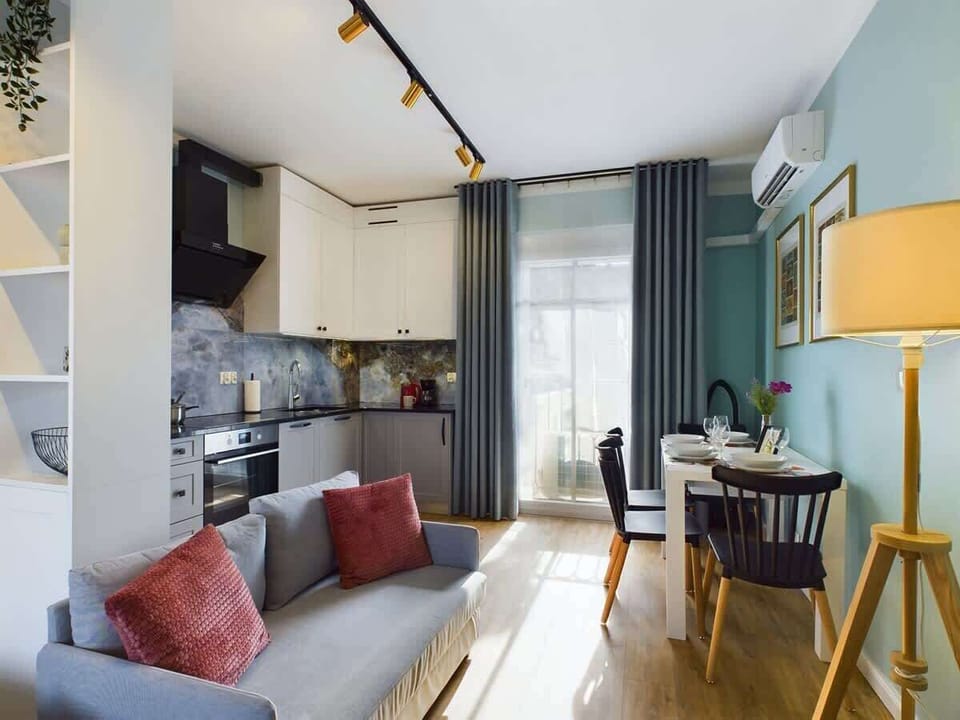  An elegant living room with sofa, bright kitchenette, and large window offers a mix of comfort and functionality.