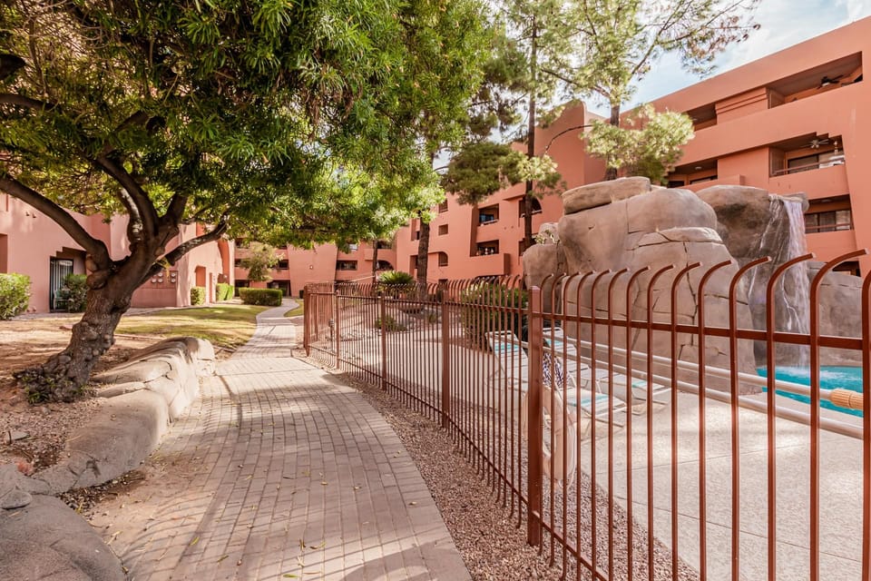 Brick-paved walkway curves beside a fenced pool with rock waterfall, surrounded by rust-colored stucco buildings.