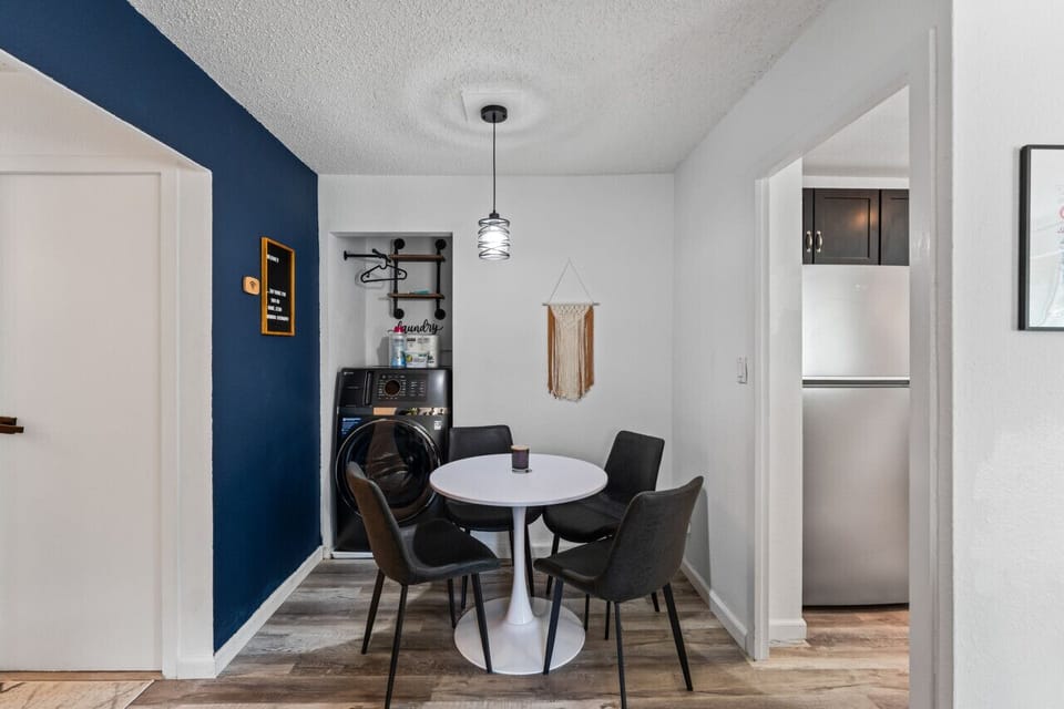 A cozy dining space with a sleek white table and black chairs sits beside a built-in laundry nook equipped with a washer, dryer, and storage shelves, all neatly tucked into the corner.