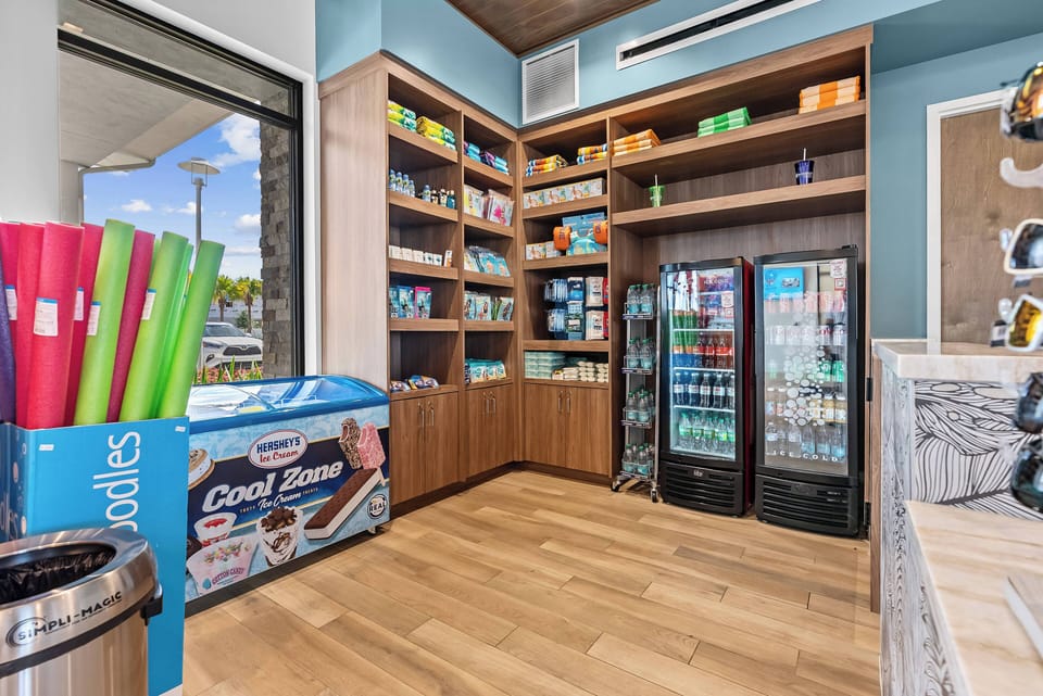 Enter a vibrant space with a sleek wooden shelf stocked with snacks and beverages, all against cool blue walls that create a refreshing atmosphere. Large windows let in natural light, making the ambiance even more inviting.