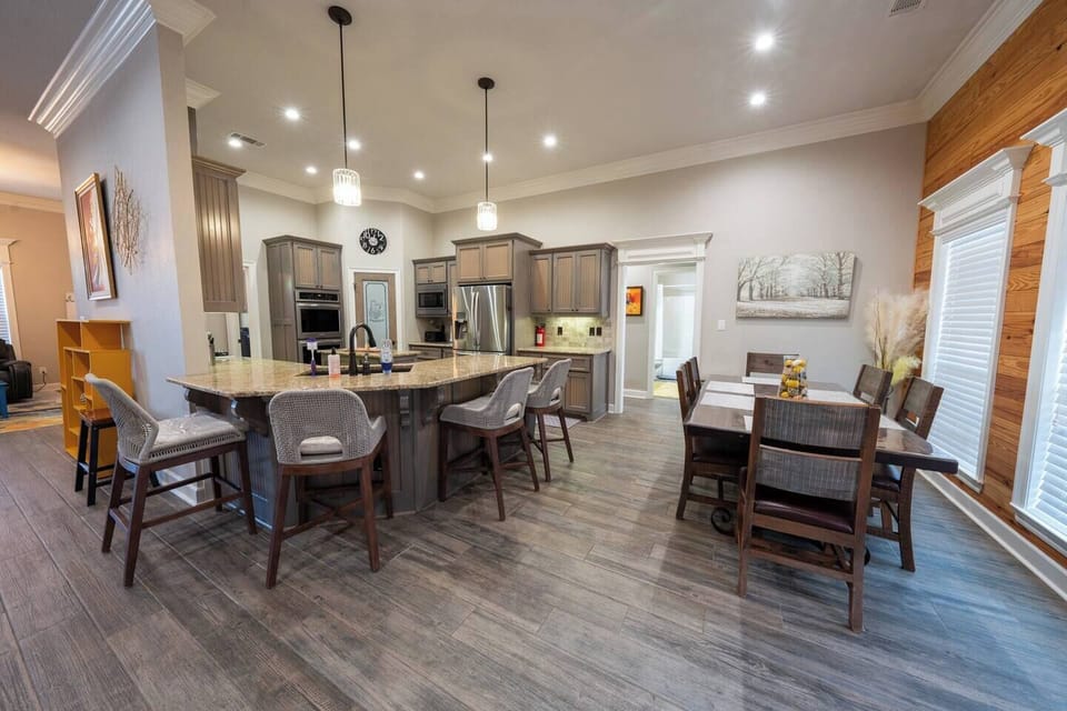 The dining area features a large, rustic wooden table with seating for six, complemented by a sleek glass vase filled with lemons as a centerpiece. The area is framed by warm, natural wood paneling on the walls, adding a cozy, cabin-like feel to the space. A bright orange door provides a pop of color and connects the dining area to the outdoor space. The large windows allow for...