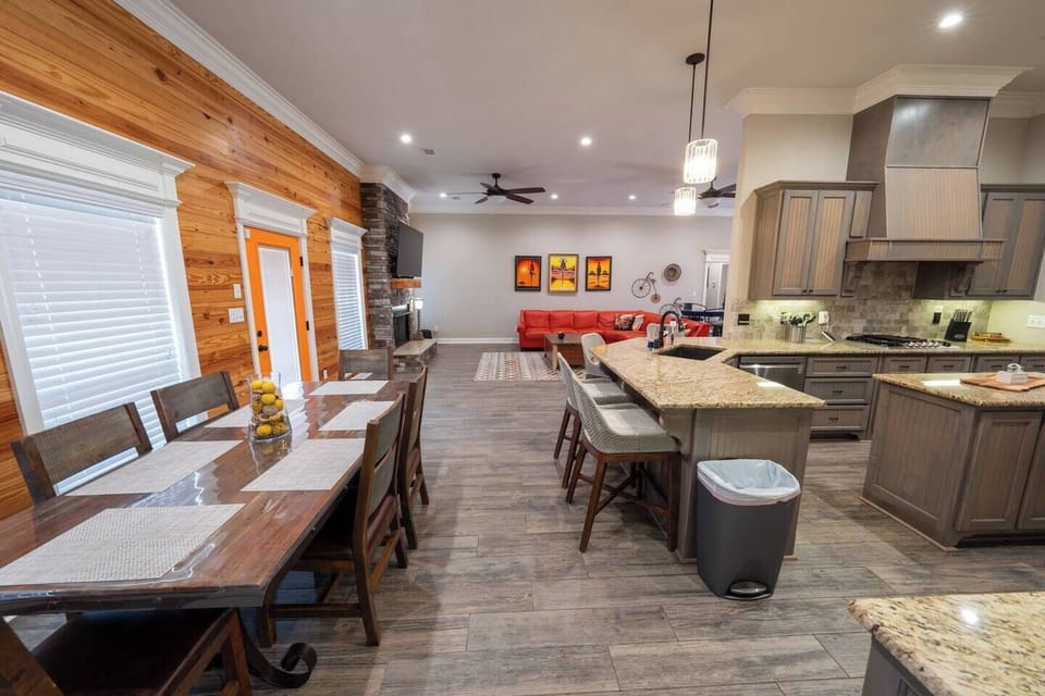 The dining area features a large, rustic wooden table with seating for six, complemented by a sleek glass vase filled with lemons as a centerpiece. The area is framed by warm, natural wood paneling on the walls, adding a cozy, cabin-like feel to the space. A bright orange door provides a pop of color and connects the dining area to the outdoor space. The large windows allow for...