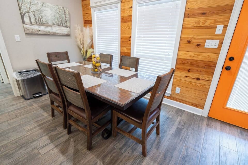 The dining area features a large, rustic wooden table with seating for six, complemented by a sleek glass vase filled with lemons as a centerpiece. The area is framed by warm, natural wood paneling on the walls, adding a cozy, cabin-like feel to the space. A bright orange door provides a pop of color and connects the dining area to the outdoor space. The large windows allow for...