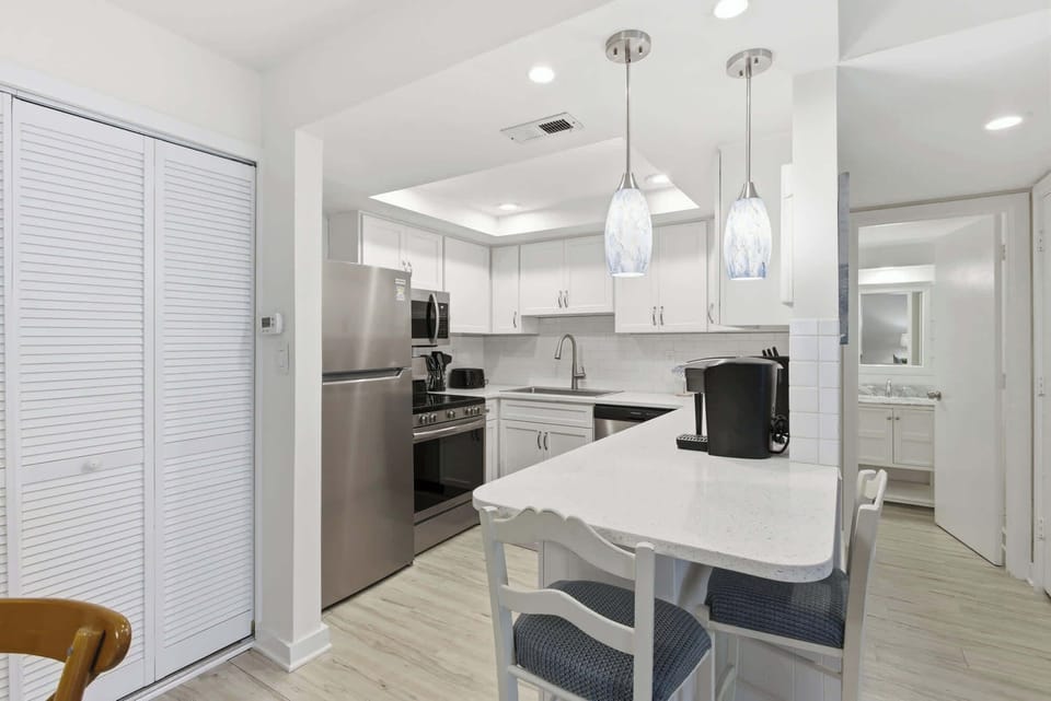White quartz counters and sleek stainless appliances sparkle under artistic pendant lights, creating a bright and airy culinary corner. This modern kitchen offers a seamless flow for brewing morning coffee or prepping a sunset dinner.