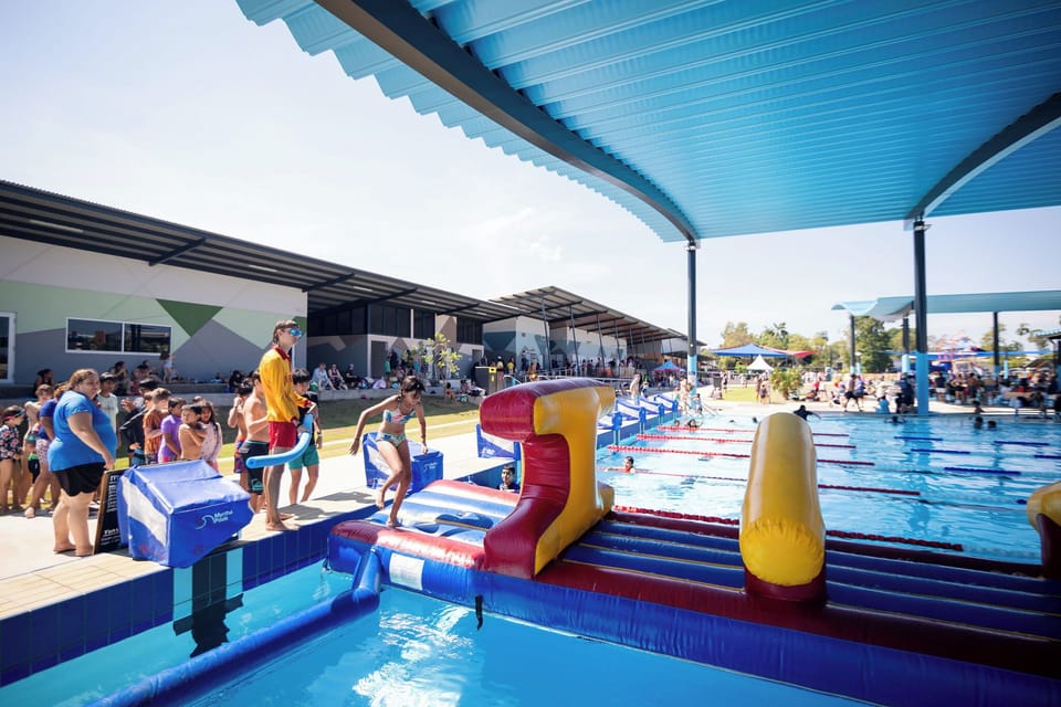 A sunny pool area with an inflatable obstacle course where kids can climb, jump, and splash. A shaded canopy over the deck gives adults a comfortable place to watch, cool off, and enjoy a relaxed day out.