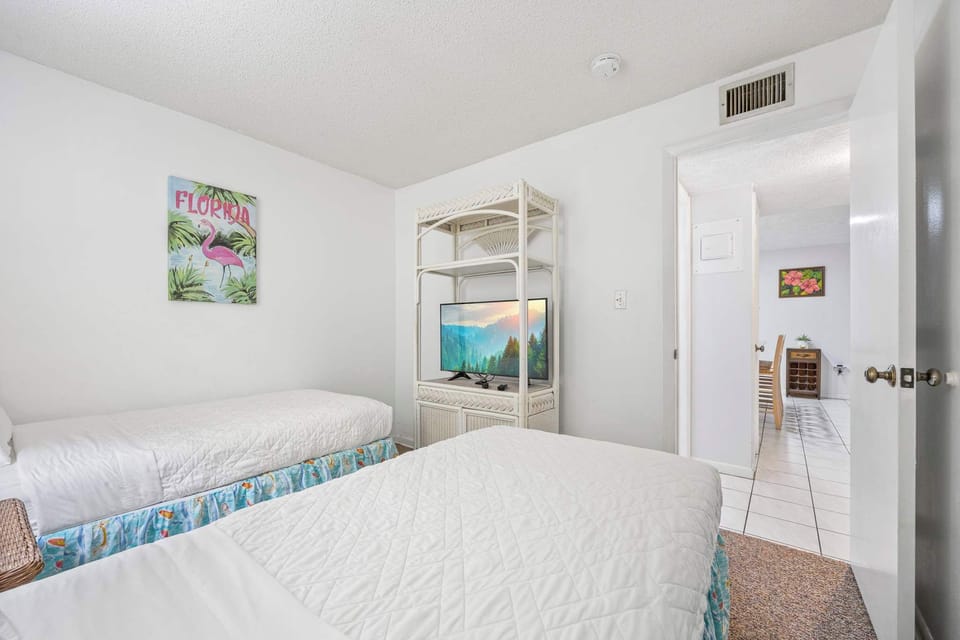 Twin beds with white linens and a television on a wicker stand in a simple, bright room.