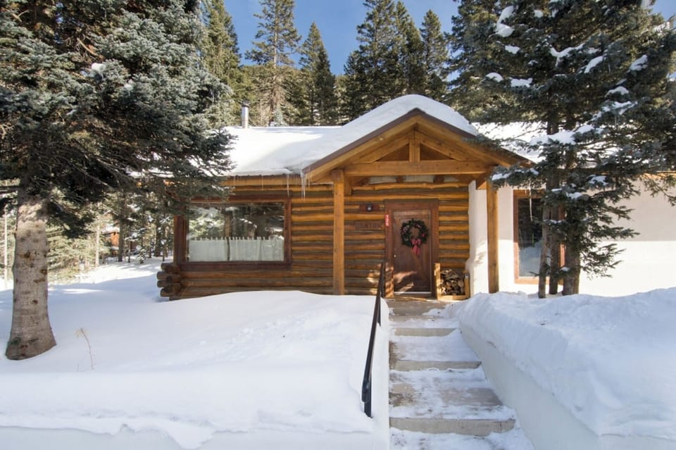 A charming log cabin nestled among snow-covered pines, featuring a welcoming entrance adorned with a festive wreath. The property offers a peaceful mountain retreat surrounded by towering evergreens and pristine winter scenery.