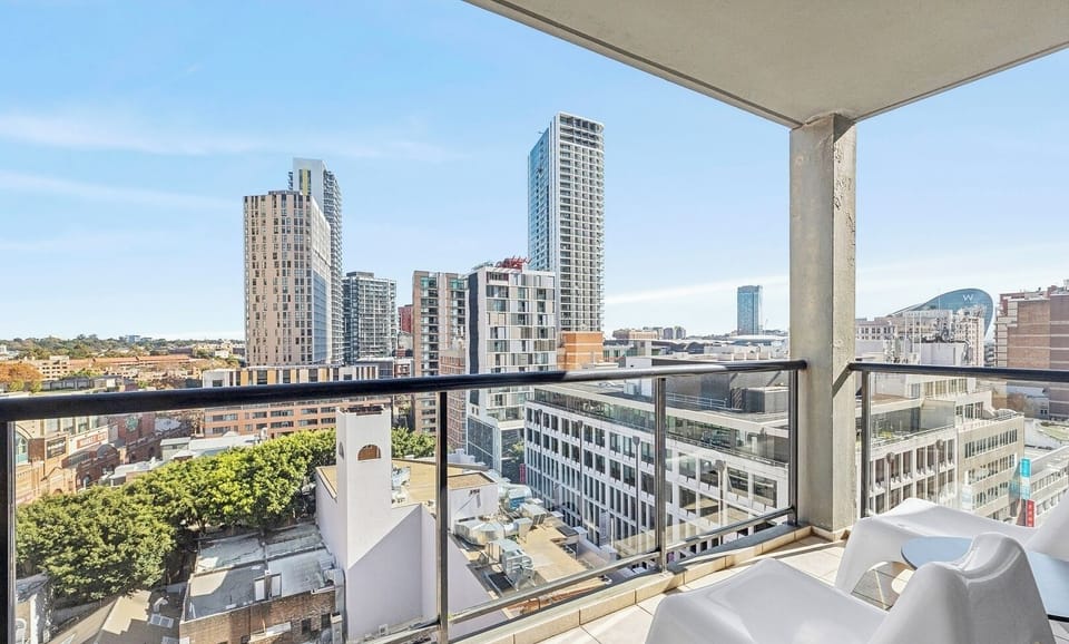 An outdoor balcony setup featuring a white chair by the glass railing, looking out onto high-rise city buildings