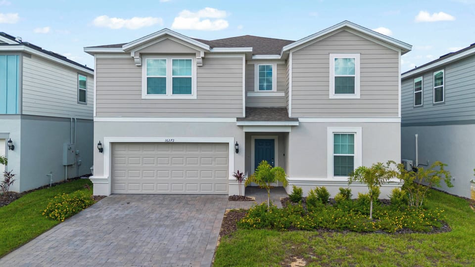 The charming exterior, featuring light gray siding and lush green landscaping, invites you in. Bright, large windows create an airy atmosphere, while the neatly paved driveway adds both convenience and curb appeal.
