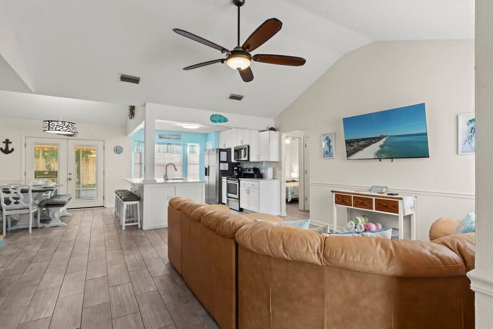 A spacious living room with tall ceilings, a comfortable leather sectional, and a beach theme, filled with natural light and an open, airy feel.