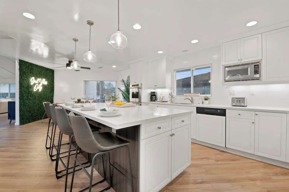 A modern white kitchen with a sleek island counter, built-in fridge, oven, microwave, dishwasher, and wine chiller. Comfy chairs line one side for casual dining, while the rest of the space provides plenty of room for meal prep.