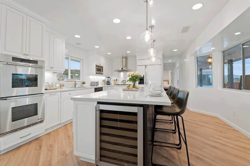 This modern white kitchen boasts a sleek island counter with built-in appliances – fridge, oven, microwave, dishwasher, and wine chiller. Comfy chairs create a cozy spot for casual meals, while the spacious layout offers plenty of room for meal prep.