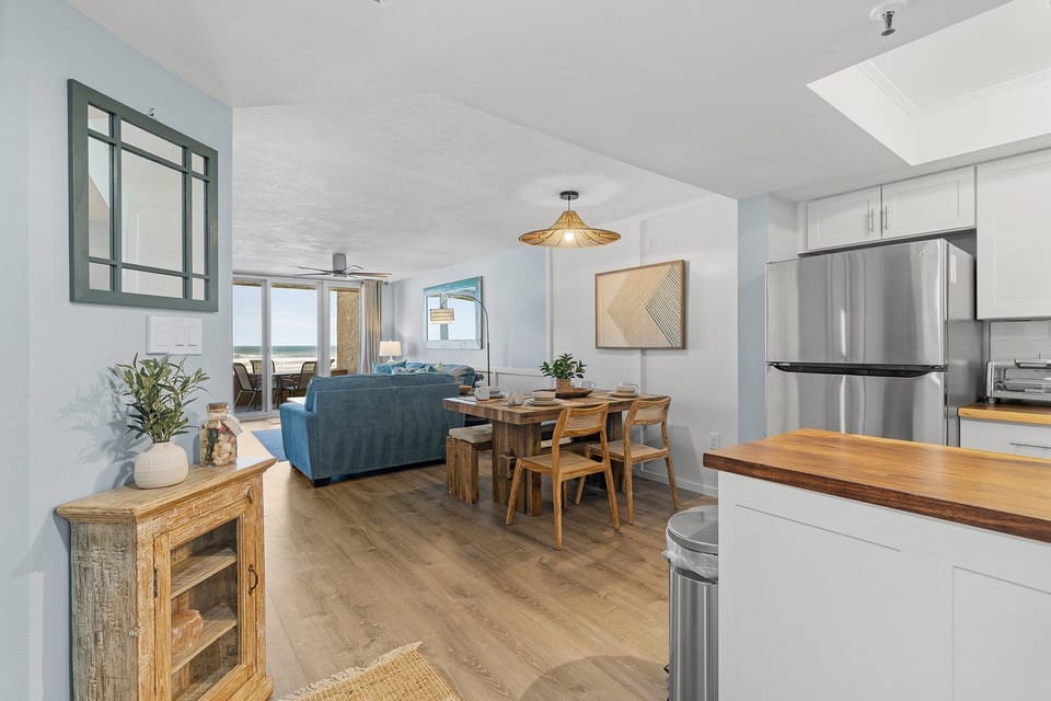 This open-concept space features a rustic dining area, a modern kitchen with stainless steel appliances, and a cozy living room. Large windows provide natural light and breathtaking ocean views, creating a warm and inviting atmosphere.
