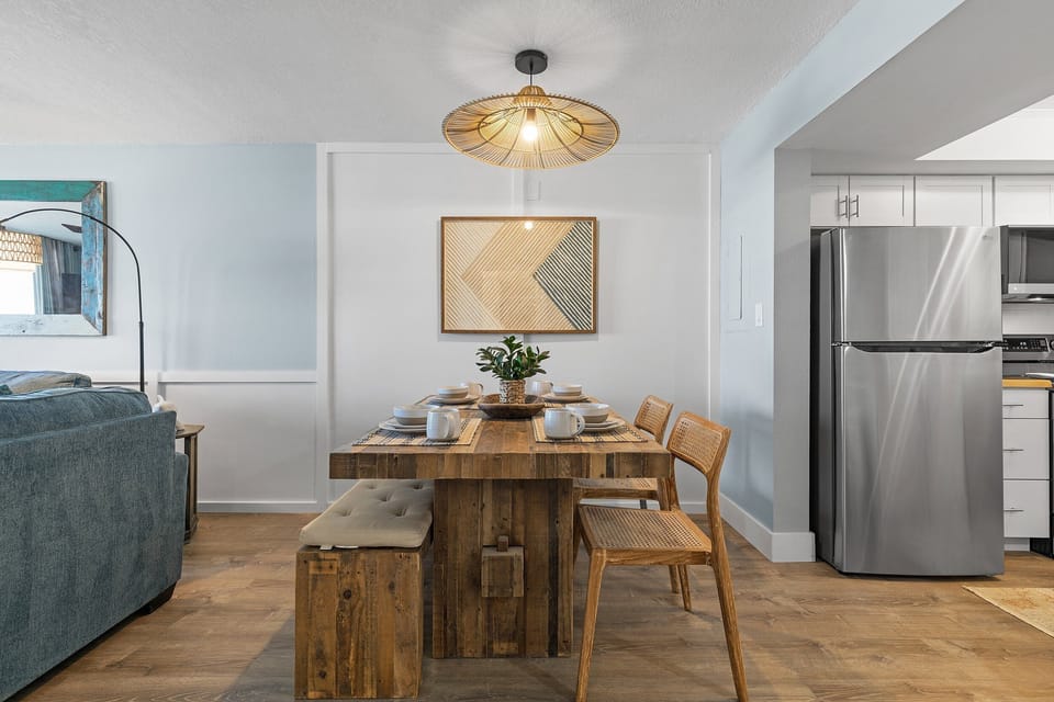 A cozy dining space featuring a rustic wooden table, cushioned bench seating, and wicker chairs. A modern light fixture and minimalist artwork add charm to the area, blending traditional and contemporary styles for a warm, inviting feel.
