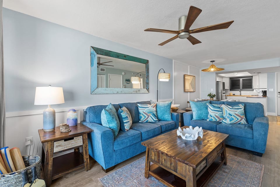 This cozy living room features vibrant blue sofas, colorful throw pillows, and a rustic wood coffee table. A large distressed mirror and coastal accents create a relaxed, beach-inspired atmosphere, perfect for unwinding by the sea.