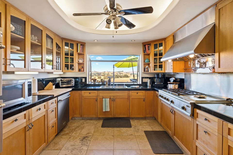 A professional chef's kitchen, featuring warm wood cabinetry, a high-end gas range with a sleek hood, and a large window overlooking the scenic hills.