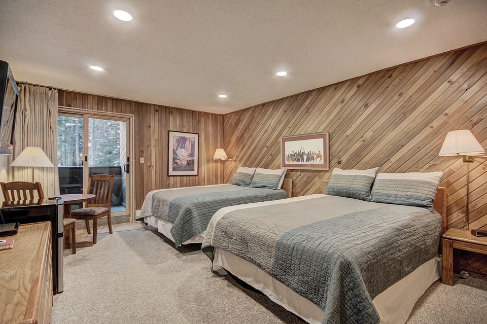 A cozy bedroom features two double beds with gray-blue bedspreads, wooden walls, a desk with a chair, and sliding glass doors leading to a balcony with a forest view.