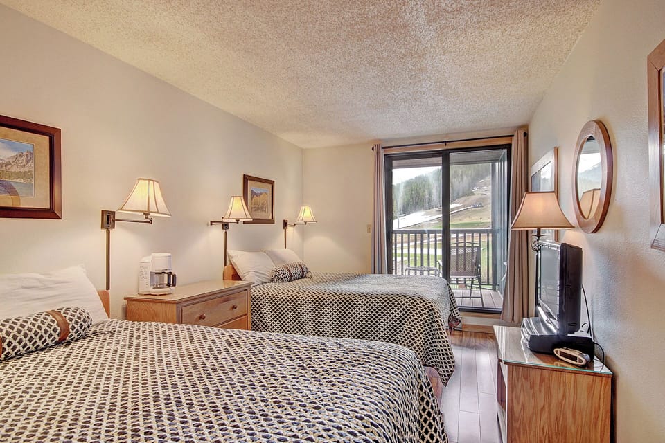 A hotel room with two double beds, bedside lamps, framed pictures, a TV, dresser, and sliding glass door leading to a balcony with an outdoor view.
