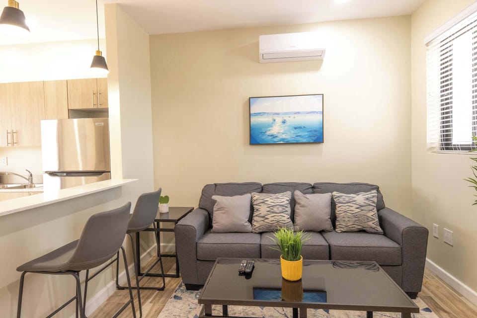 A cozy, modern living room with a sleek couch, plants, and a kitchen area in the background.
