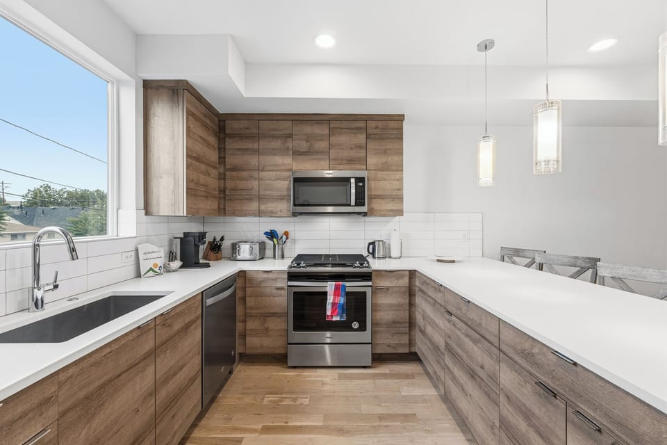 Step into your modern kitchen with warm wood cabinetry, bright white countertops, and stainless appliances ready for your culinary adventures. Sunlight streams through the window, illuminating your cooking space.