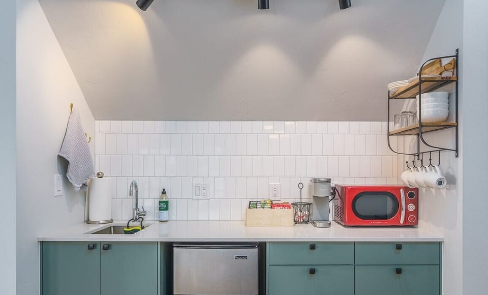 - Well lit kitchenette with green cabinetry, white tile backsplash, and sleek black hardware
- Fully equipped with microwave, mini fridge, sink, Keurig, mugs, and open shelving
- Under cabinet lighting and wall track lights create a stylish ambiance