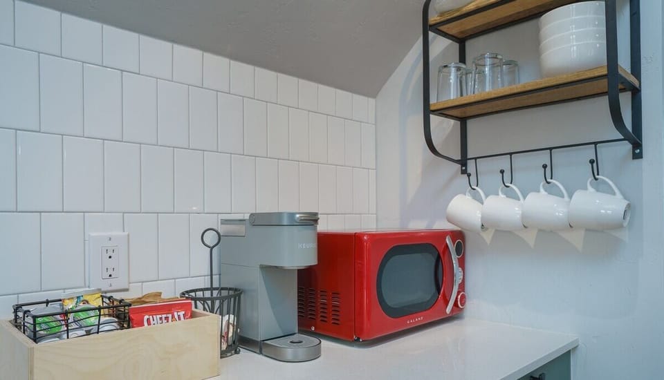 - Kitchenette essentials include a bold red microwave, Keurig, mugs, and tea station
- Hanging mugs and glassware are neatly displayed on modern open shelving
- Sleek white countertops and vertical tile create a crisp clean workspace