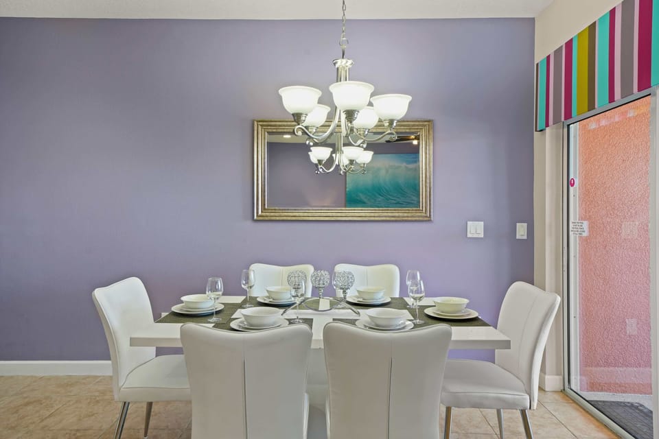 - Elegant dining area featuring a modern white table set with matching chairs and glassware - Chandelier adds a touch of sophistication, complementing the soft purple accent wall - Mirror and colorful striped valance bring a vibrant, stylish feel