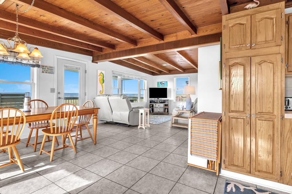 Cozy living room with wood-beam ceiling, recliners, wall TV, ocean views, large windows, deck access, seahorse art, coral rug, and tile floors.