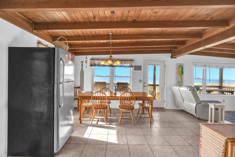 Dining area with wood table, chandelier, ocean views, deck door, wood-beam ceiling, coastal curtains, seahorse art, and beach-facing windows.
