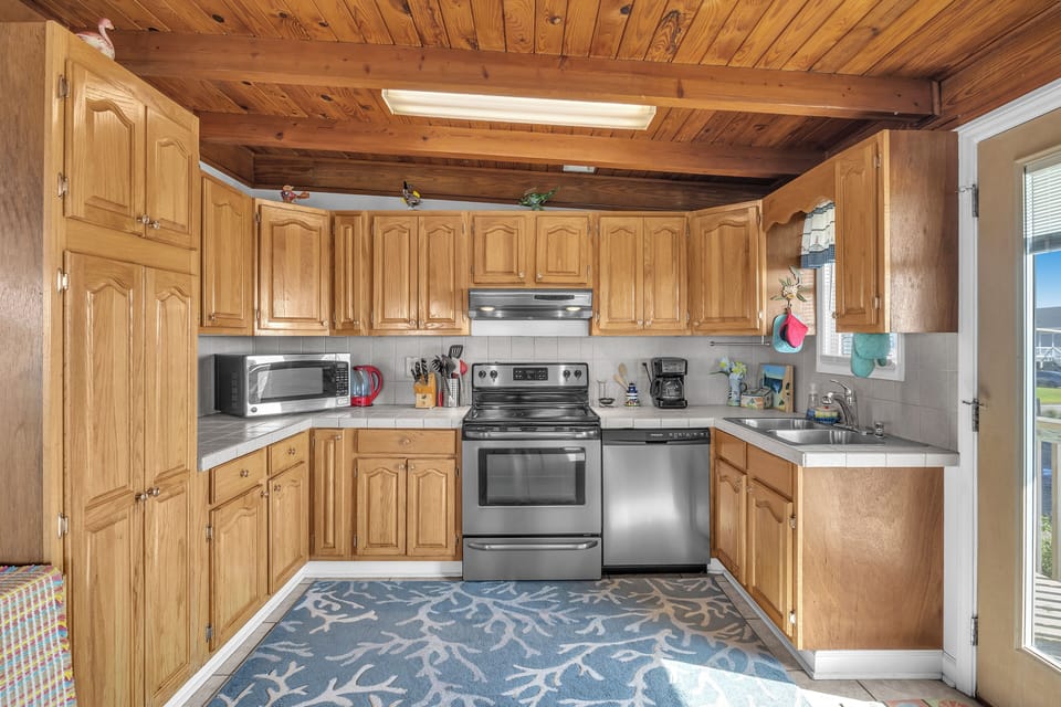 Cozy kitchen with wood cabinets, stainless appliances, tile counters, double sink, microwave, coffee maker, wood ceiling beams, coral rug, and ocean-view door.