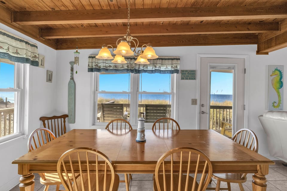 Dining area with wood table, chandelier, ocean views, deck door, wood-beam ceiling, coastal curtains, seahorse art, and beach-facing windows.