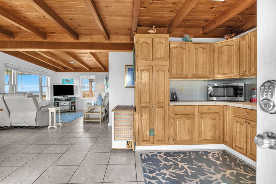 Cozy kitchen with wood cabinets, stainless appliances, tile counters, double sink, microwave, coffee maker, wood ceiling beams, coral rug, and ocean-view door.