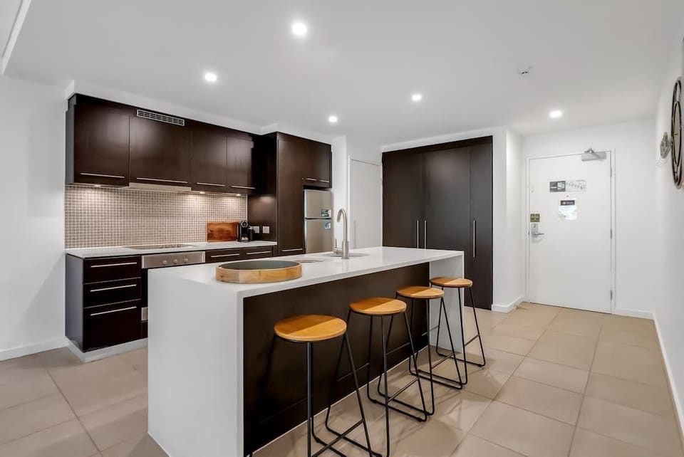 The sleek island bench offers a casual spot to enjoy drinks and platters, blending seamlessly with the kitchen’s modern dark cabinetry and stone finishes.