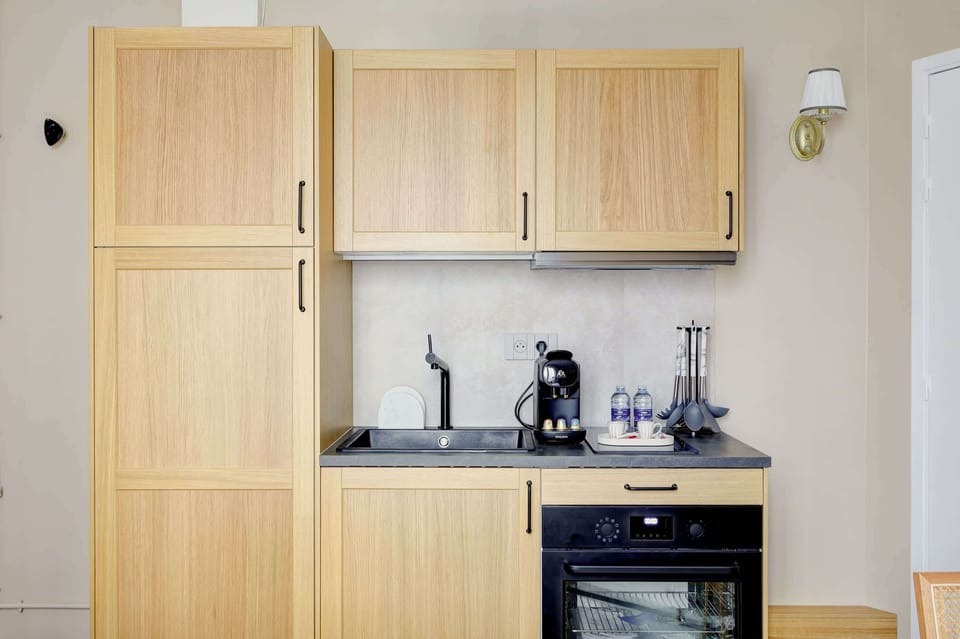 La kitchenette moderne de ce studio, avec ses armoires en bois clair et ses finitions noires élégantes, est à la fois fonctionnelle et design. Équipée d'un four, d'une cafetière et de rangements optimisés, elle s'intègre parfaitement dans l'espace. 