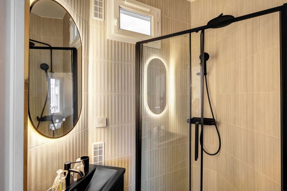 Salle de bain élégante avec une faïence couleur amande accompagné d'une robinetterie noir, doté d'un meuble vasque avec en partie basse du rangement, ainsi que d'un miroir rétro-éclairé et anti-buée.