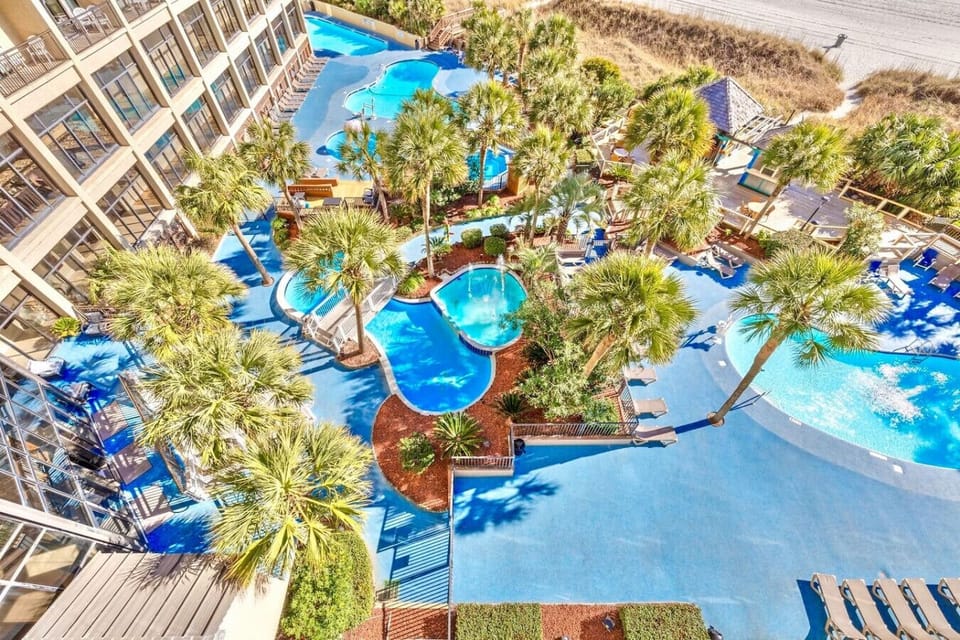 Private balcony view overlooking tropical resort pools, swaying palms, and the oceanfront setting at Beach Cove Resort in North Myrtle Beach.