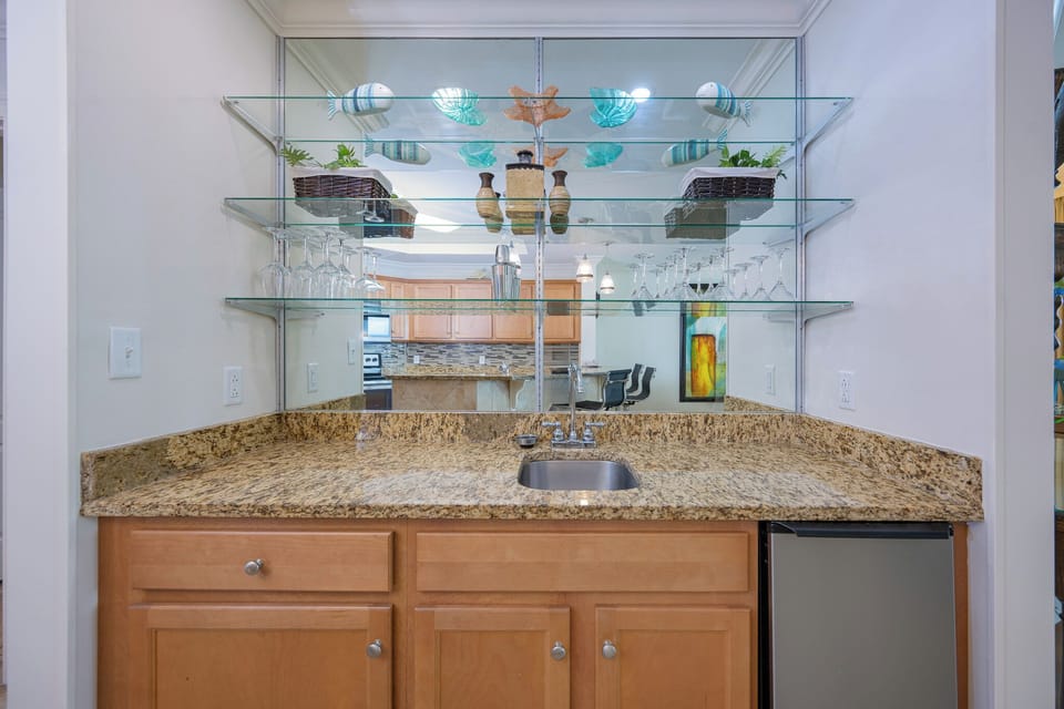 Wet bar area with granite countertops, blender, ice-maker, and a view of the cozy living area and balcony beyond