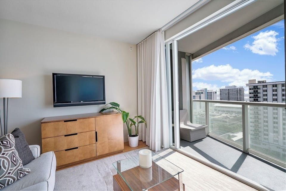 Modern living room with a wall-mounted TV, wooden dresser, sofa, and glass coffee table. A sliding glass door leads to a balcony overlooking partial ocean views and intracoastal waterway.