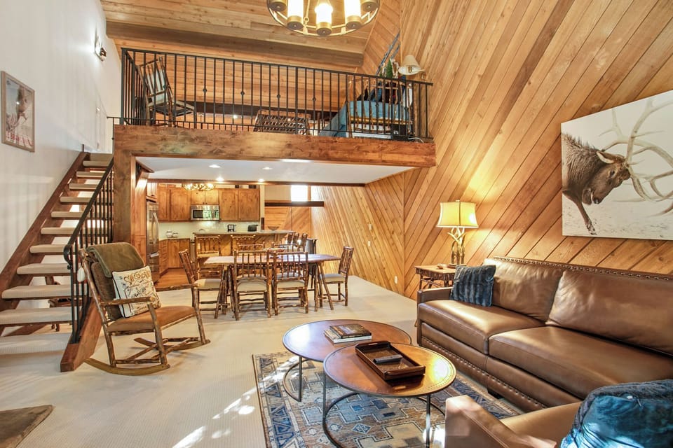 A living room with wood-paneled walls, a loft, leather couch, coffee tables, armchairs, and dining area. Stairs lead to the loft. Decor includes a cowboy picture and a bull painting.