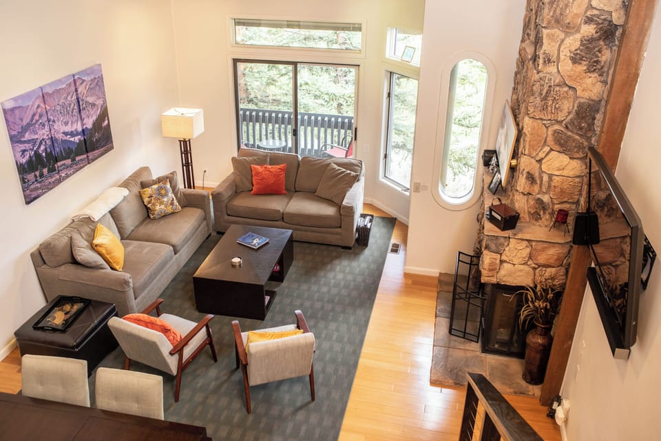 A modern living room with two gray couches, two gray armchairs, a central coffee table, a large stone fireplace, and large windows overlooking a forest. The decor includes a painting and multiple cushions.
