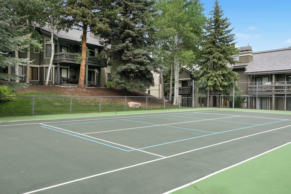 A tennis court with clearly defined lines in front of two-story residential buildings surrounded by tall trees and greenery.
