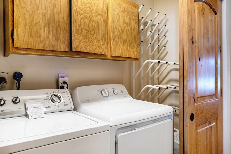 A laundry room with a washer and dryer beneath wooden cabinets, a wooden door on the right, and a wall-mounted drying rack.