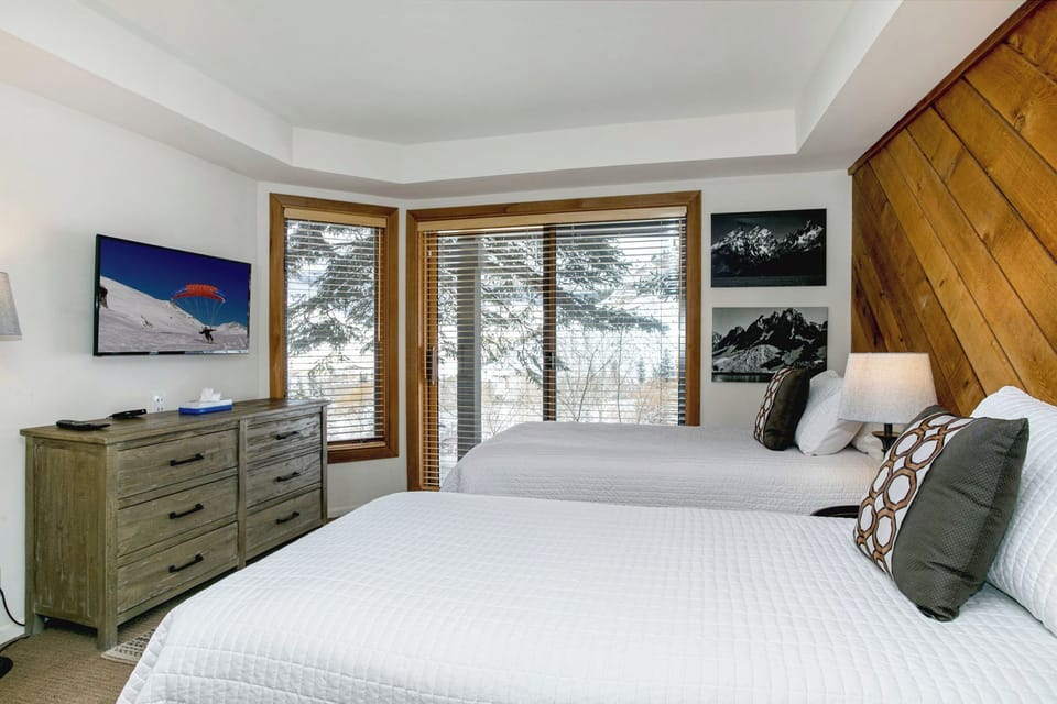 A bedroom with two white beds, a dresser with a TV, a window with blinds, and pictures on the walls.