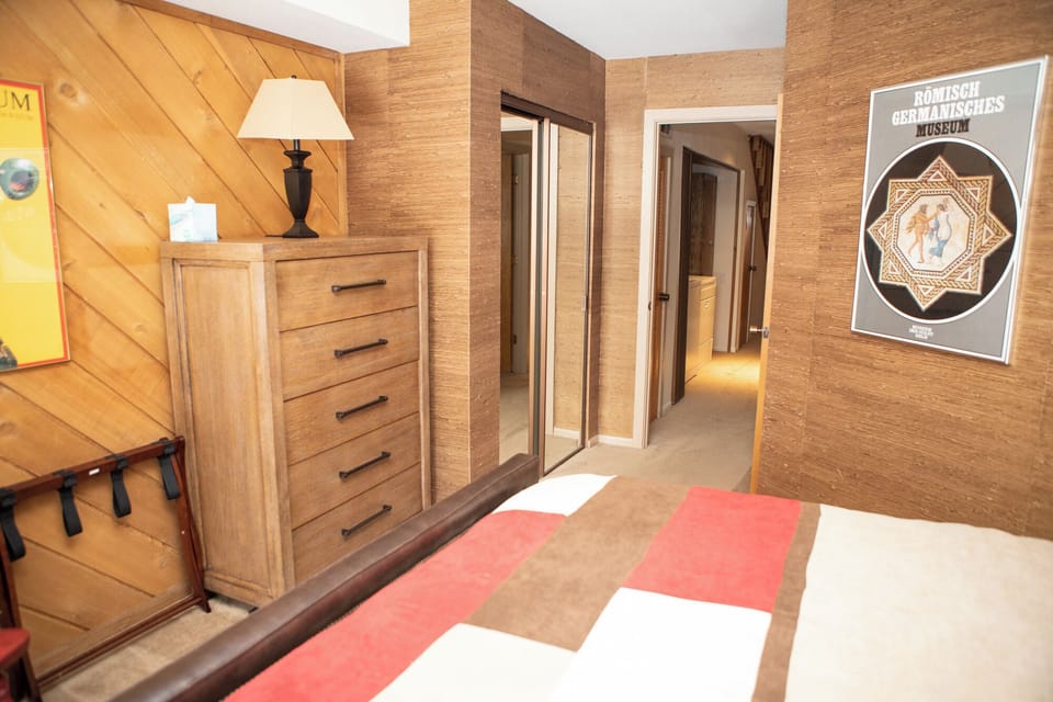 A bedroom with a wooden dresser, a bed with a multicolored blanket, a lamp, a poster on the wall, and open doors leading to other rooms.