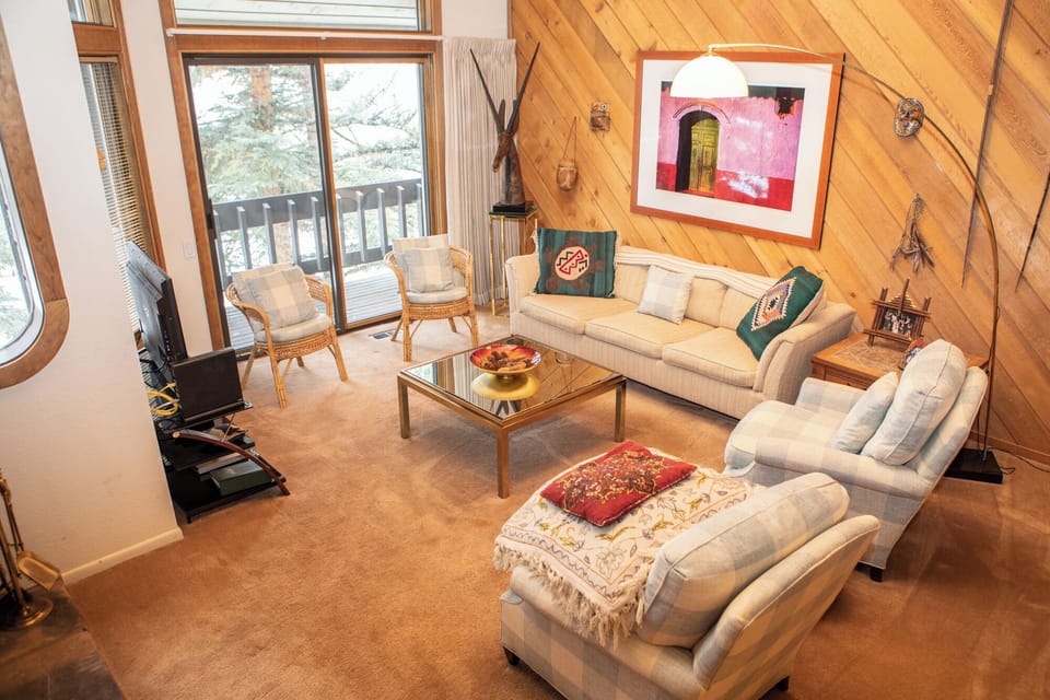 A cozy living room features beige sofas, a glass coffee table, and plaid armchairs. Art decorates a wood-paneled wall, while a sliding glass door leads to a snow-covered balcony outside.