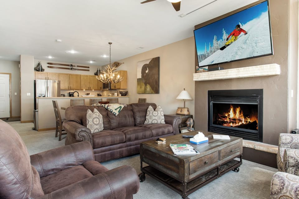Cozy living room with brown sofas, a fireplace, wall-mounted TV showing a skier, and a coffee table. The kitchen with wooden cabinets is visible in the background. Decorative items adorn the space.