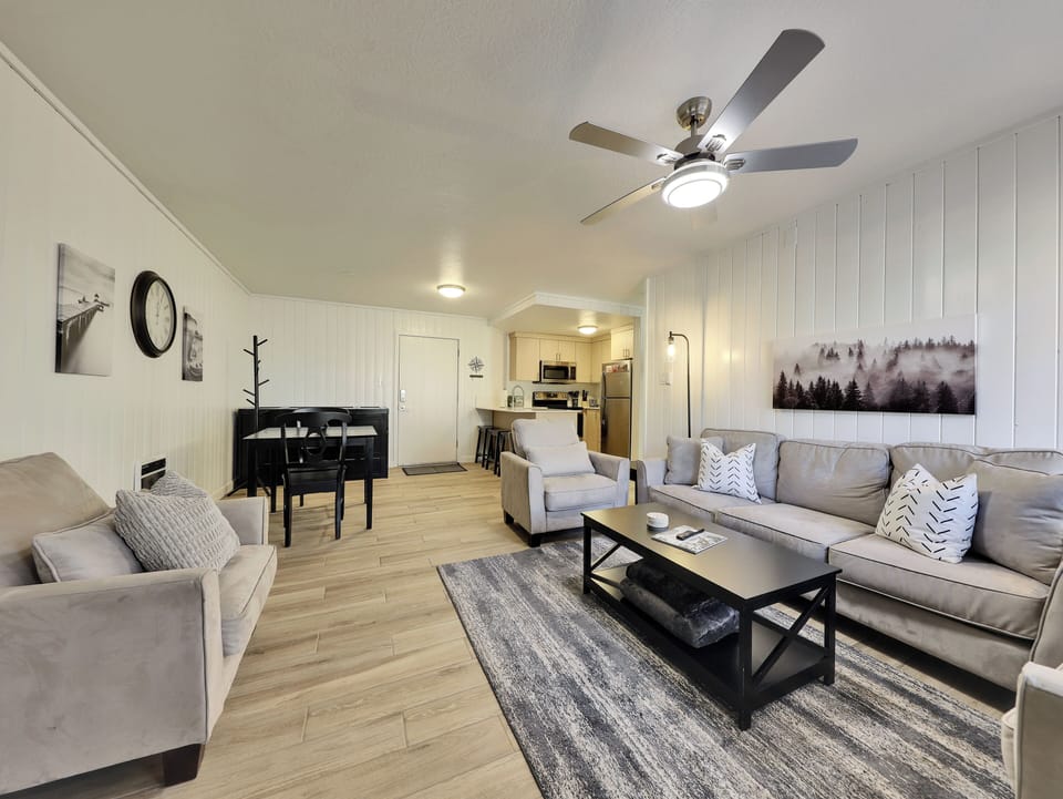 View of the newly updated, light, and airy living room and dining area, featuring a 58" smart TV, electric fireplace, ceiling fan, and comfortable sofa and armchairs.