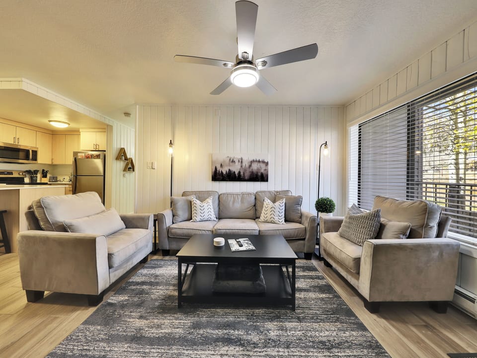 View of the newly updated, light, and airy living room featuring a 58" smart TV, electric fireplace, ceiling fan, and comfortable sofa and armchairs. A glass door leads to the private walk-out balcony