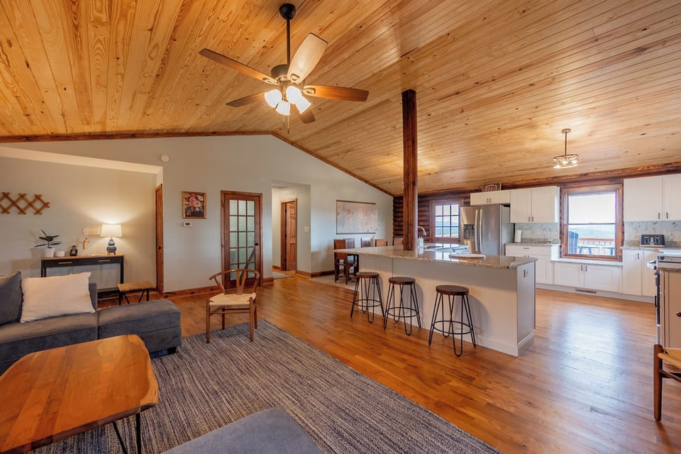 This inviting view captures the seamless flow between the living area and kitchen at Valley View Getaway. The warm wooden ceilings and open layout create a spacious feel, perfect for gatherings. The kitchen features a large island with bar seating, while the cozy living space offers comfortable seating and tasteful decor. Natural light pours in through the windows, highlighting...