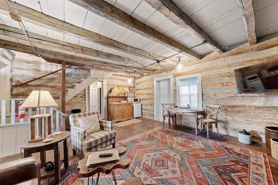A sweeping view connects the rustic living area, dining space, kitchenette, staircase, and exposed log-and-beam architecture.